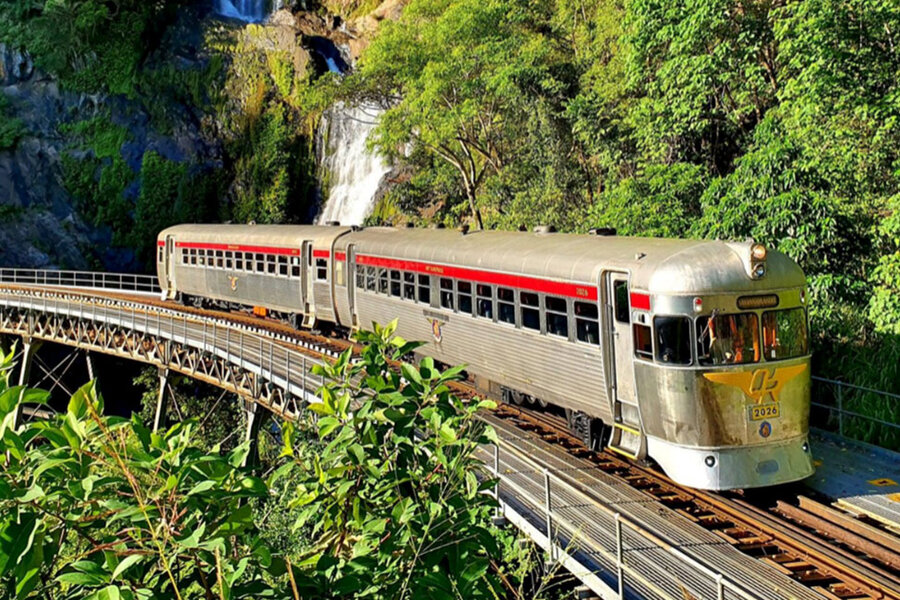 Vintage silver train with red accents crossing a bridge near waterfall and lush greenery on the Captain Cook Highwa