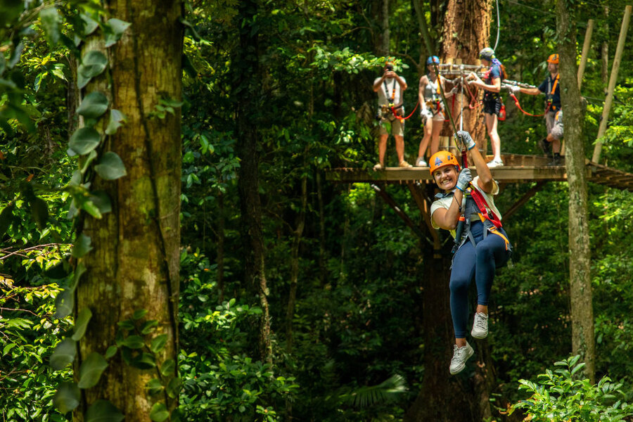 Person ziplining joyfully through a lush green forest while others wait on a wooden platform, wearing helmets and harnesses