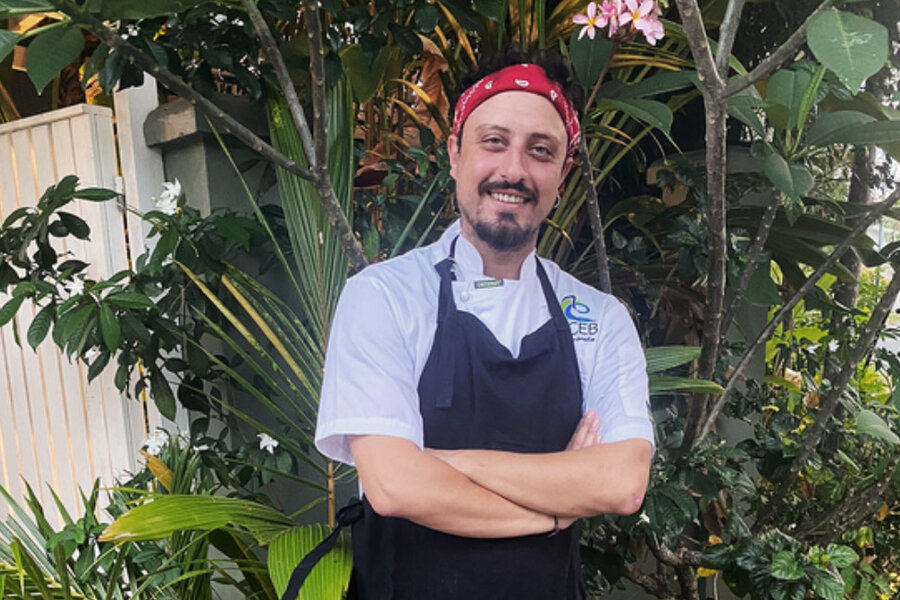 Chef Nicola Musacchio smiles with arms crossed, wearing a red bandana and black apron, standing in a lush garden with tropical plants and a white fence.