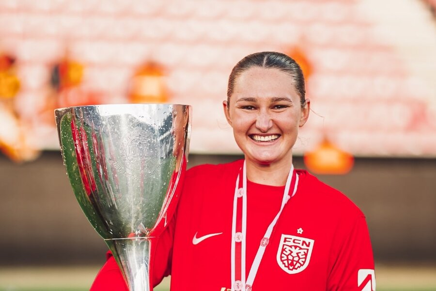 Wini Heatley smiling in a red FCN jersey while holding a large silver trophy, with a blurred stadium background suggesting celebration
