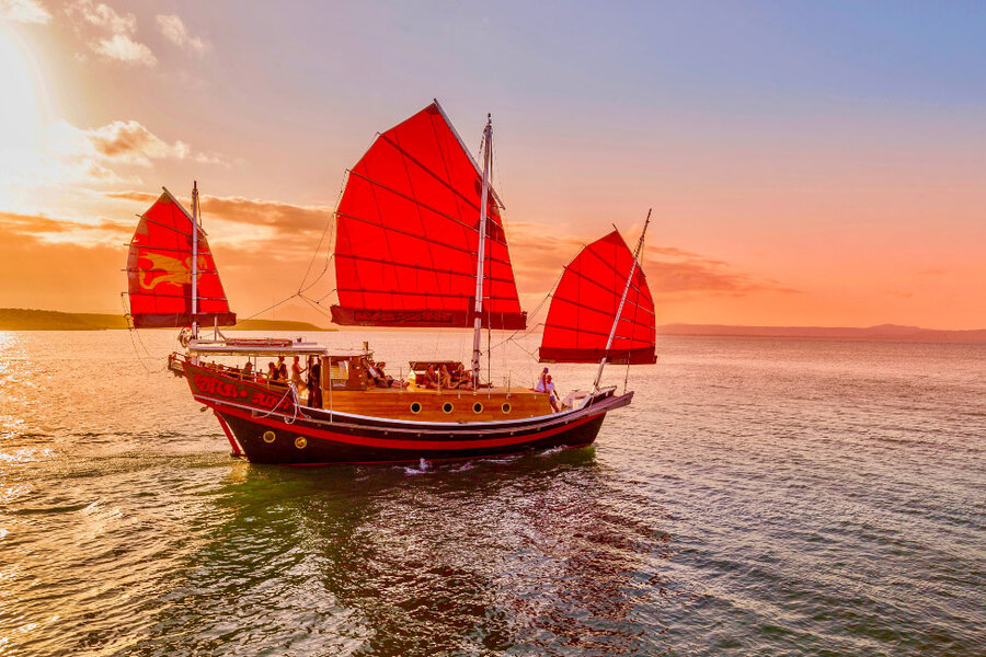Traditional sailboat with red sails gliding on calm water at sunset in Port Douglas, with orange and pink sky