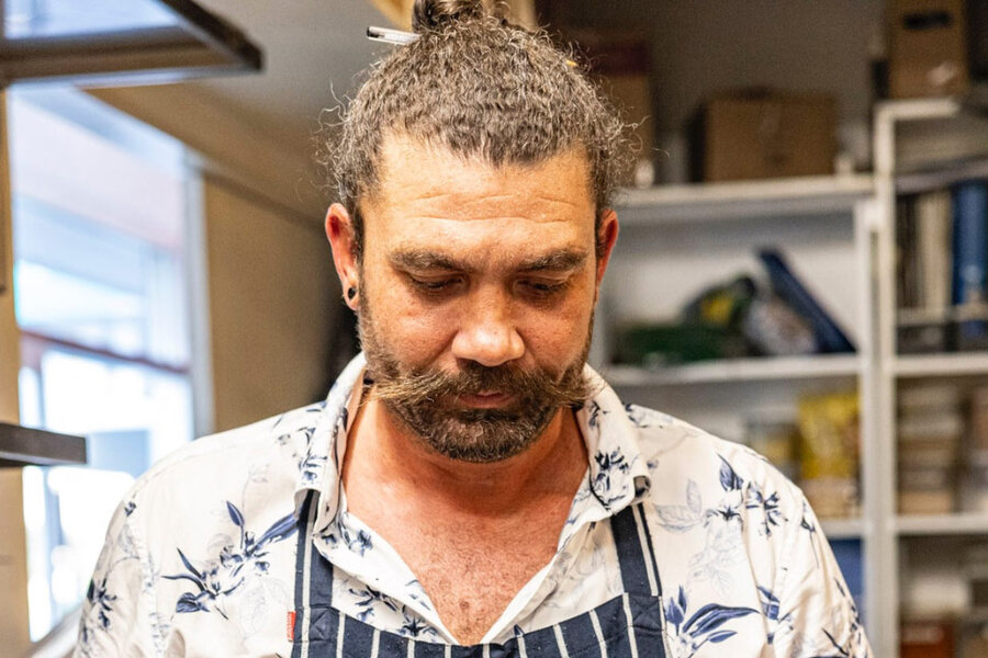 Evan Seaward, a bearded man with a bun and patterned shirt, wearing an apron, stands focused in a kitchen with shelves full of items behind him