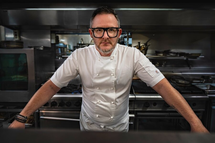 Chef Spencer Patrick in a white uniform and glasses stands confidently in a professional kitchen, surrounded by stainless steel equipment.