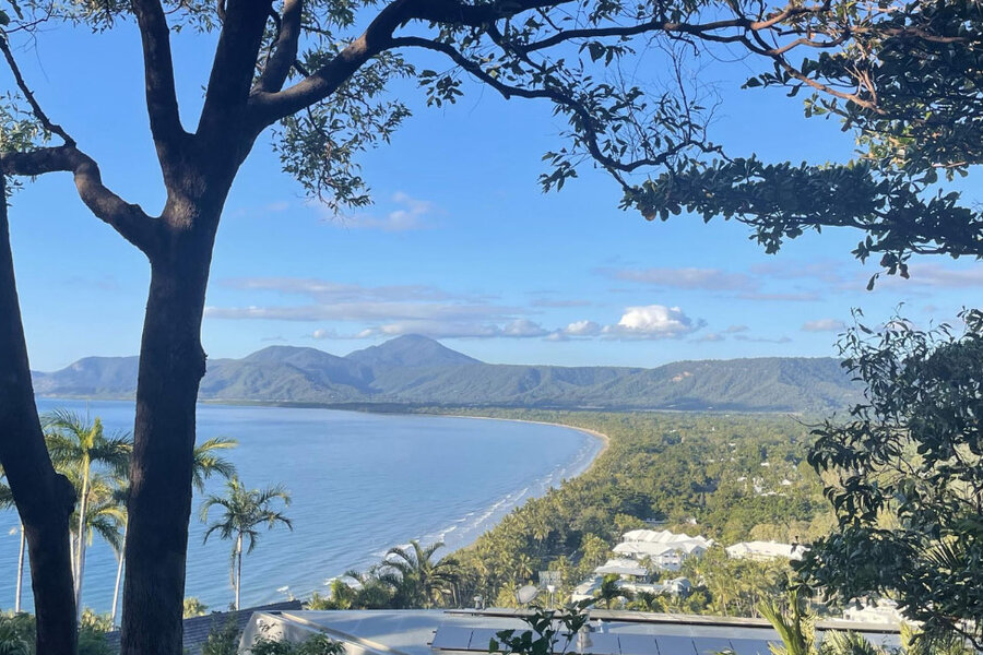 Scenic view of Four Mile Beach framed by tree branches, with curved shoreline, lush greenery, and distant mountains.