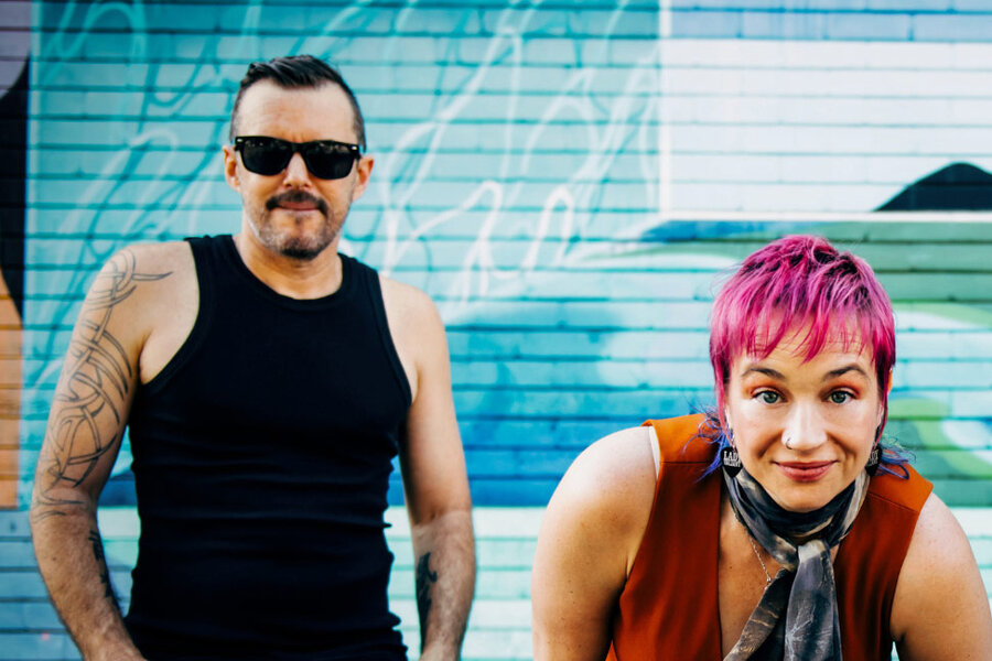 Glenna and Marty pose in front of a colorful mural; he wears a black tank top and sunglasses, she smiles warmly with pink hair