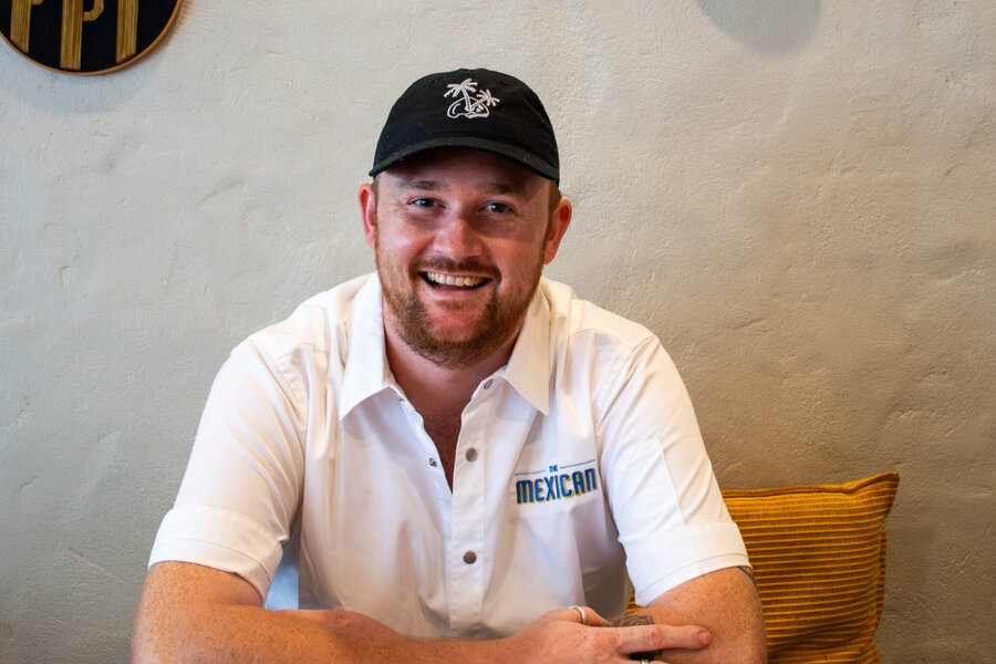 Dylan Boyle at The Mexican, smiling in a white shirt and black cap, seated against a textured wall with a yellow cushion, giving a relaxed vibe.
