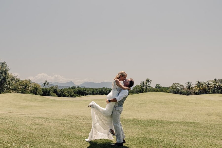 SCOTT + SONJA – Captured by RebeccaWadePhotography, a couple in wedding attire embraces on a sunny grassy field; the groom lifts the smiling bride under a clear sky with distant trees
