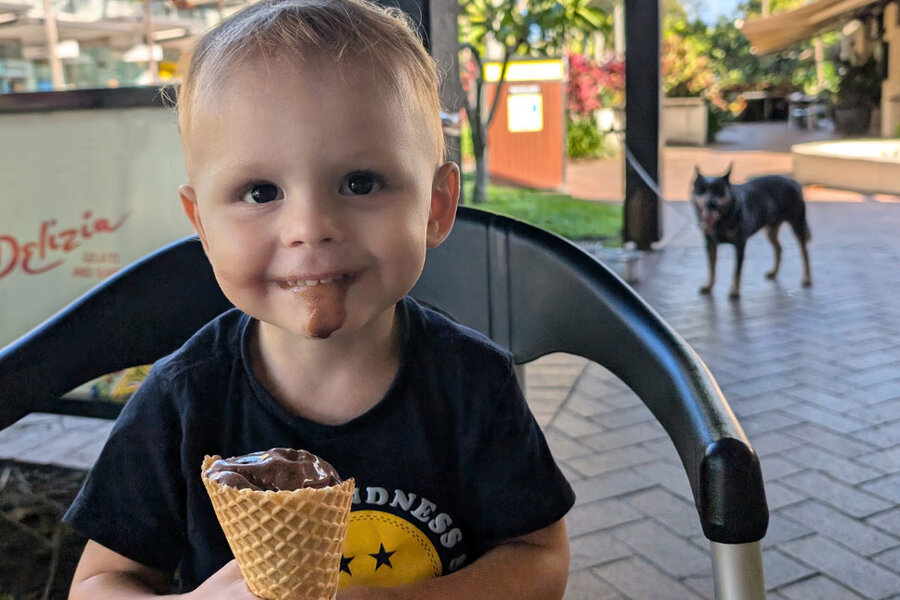 Smiling toddler with chocolate ice cream on their face holding a cone, sitting outdoors on a patio with a dog in the background creating a joyful scene